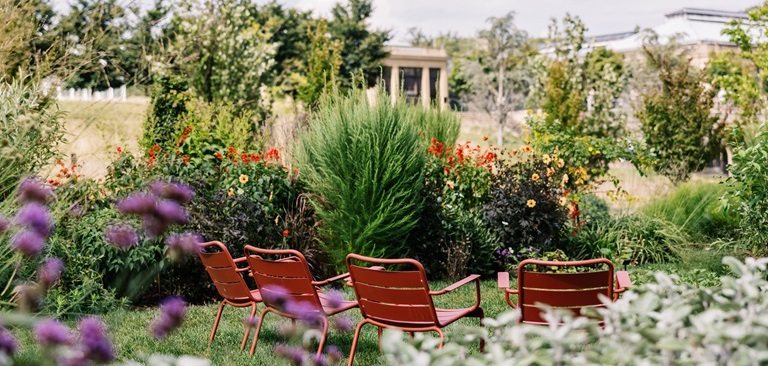 Four empty copper colored chairs arranged in a line in a lush garden.