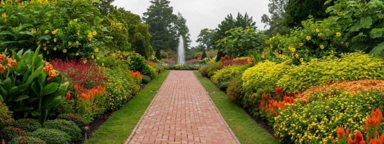 View down the length of a brick walk bordered by colorful beds of late summer flowers, leading to a central fountain in the distance.