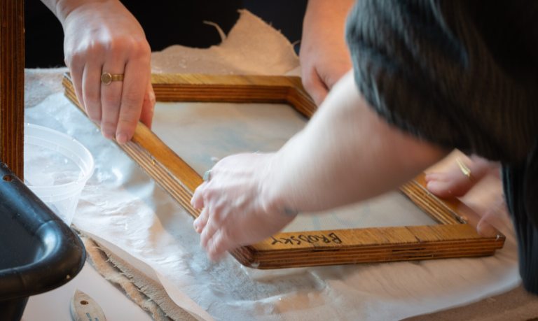 Two people holding a wood frame and making paper.