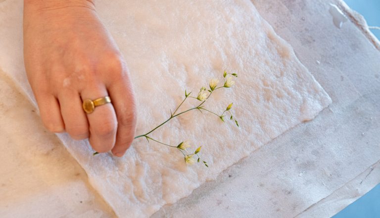 A persons hand placing a delicate flower on top of a white linen type paper.