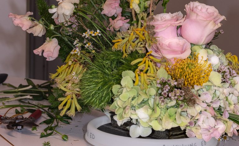 A floral design in process, featuring pink roses, goldenrod accents, and other floral material, in a shallow white vase.