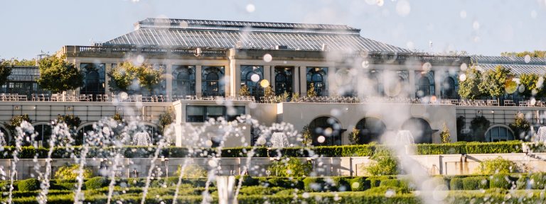 Closeup of fountains splashing in the foreground, against the backdrop of a large conservatory complex.