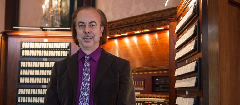 a person dressed in a dark suit, purple shirt, and tie, faces the camera with his back to a large organ console