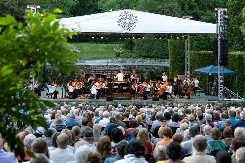 A crowd sits in front of a stage in the summer time. A symphony orchestra is on the stage.