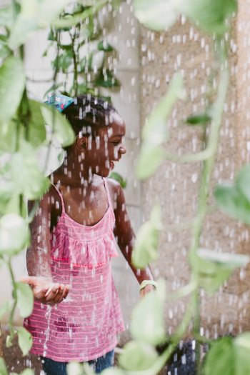 A young child is seen through a curtain of water among hanging green vines
