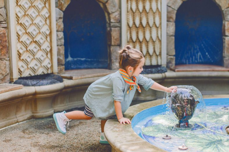 A small child stands on one foot as they reach their hand on to a stone water fountain