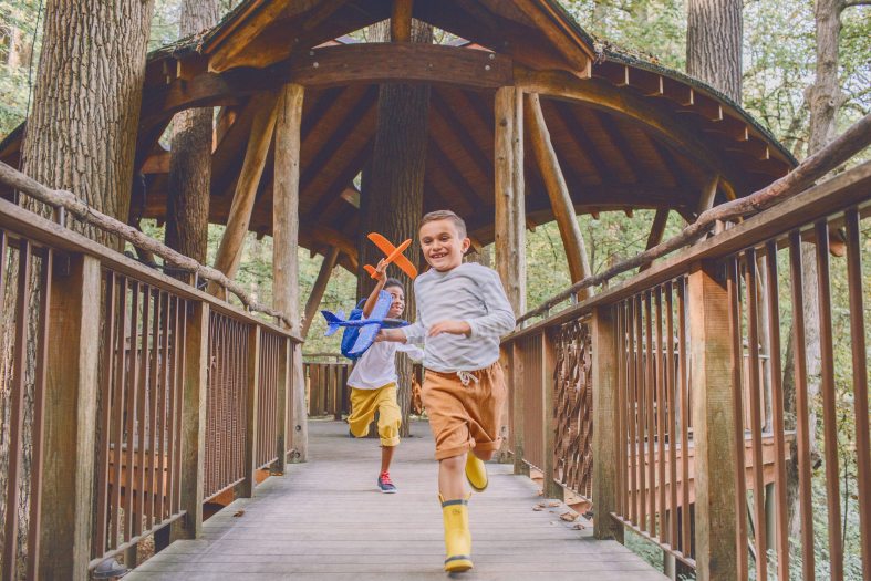 Two children holding toy airplanes run along a wooden walkway out of a wooden treehouse