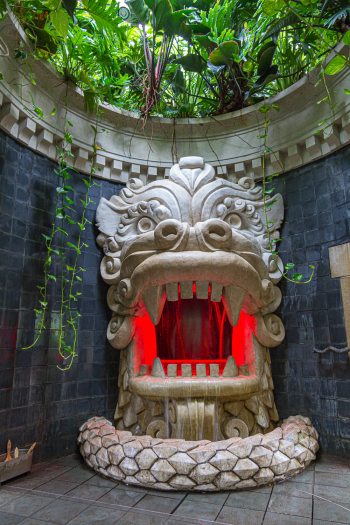 A stone dragon sculpture with an open red mouth and water feature sits in a circular room below green plants