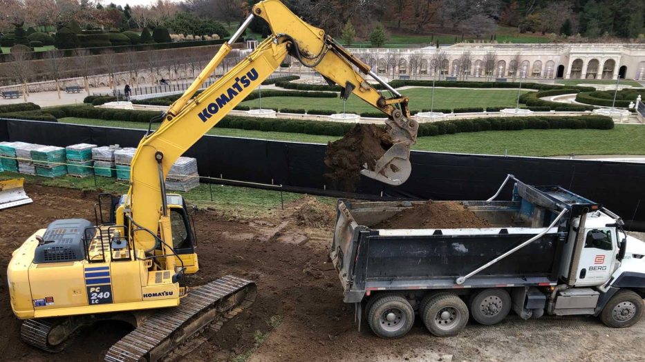 Yellow digger empties soil into a dump truck, with fountain garden in the background.