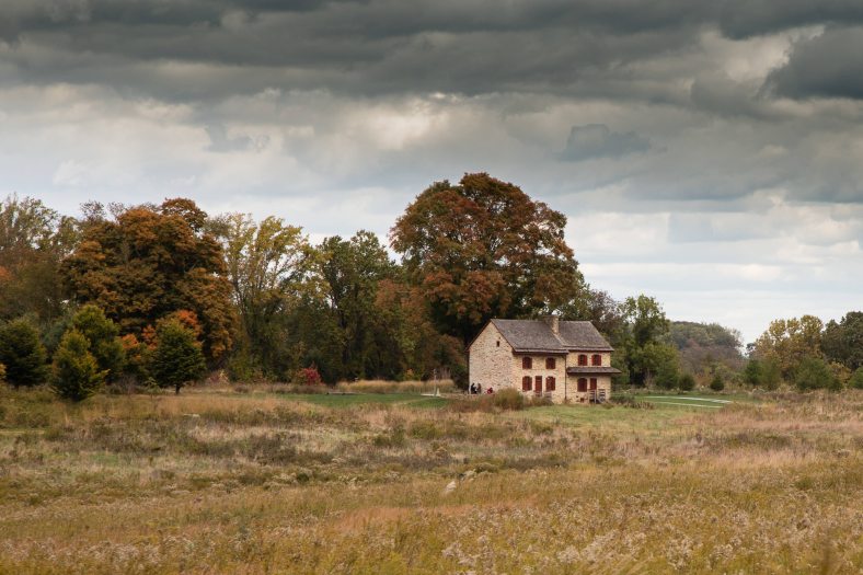 a brick 2-story farmhouse with brown roof and red shutters with field in front and trees in background, against a cloudy sky