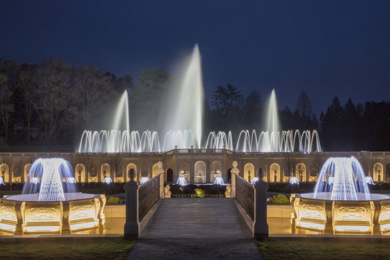 Fountains shoots water in the air illuminated by white lights across carved white stone structures