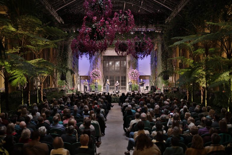 A large hall full of people looks on to a stage performance under hanging baskets of pink orchids at night in a conservatory