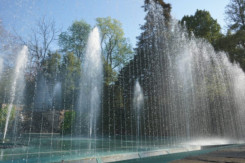 Fountains shoot into the air from a stage during a daytime fountain show