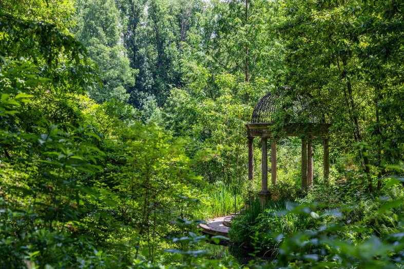 A stone gazebo sits beside a small lake tucked into a forest of bright green trees