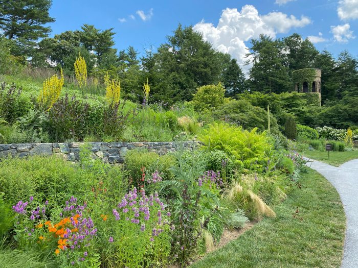 A large area of green plants and colorful flower beds with a small pond and stone tower in the distance