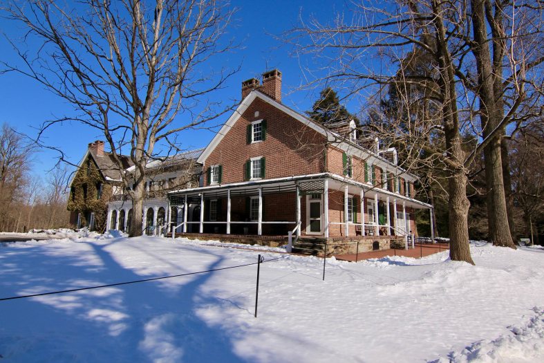 A brick and stone house with a wraparound porch is seen on an angle surrounded by snow-covered ground and trees