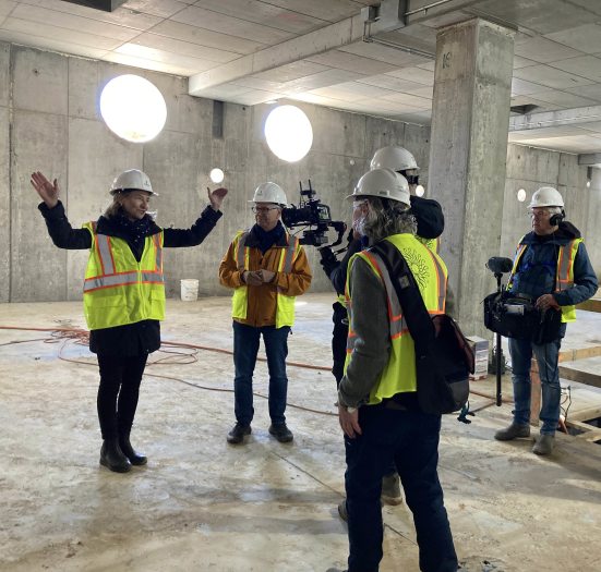 5 people in hard hats and reflective safety vests gather in a well-lit concrete basement; one person speaks and gestures, another looks on, and 3 carry recording equipment.