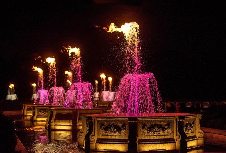 A row of octagonal stone basins at night, with fountains in basketweave formation, lit up in fuchsia and topped by fire.