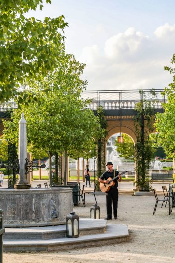 A man plays a guitar standing among an area with fountains, tables, chairs, and trees