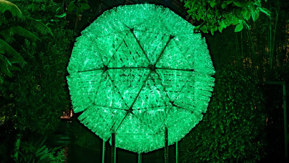 a large sphere composed of plastic bottles, lit in green against a backdrop of green garden plants