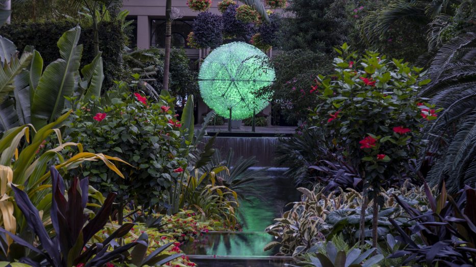 a geodesic dome of plastic bottles, lit with green light, sits amid the greenery and waterfall of a Conservatory garden 