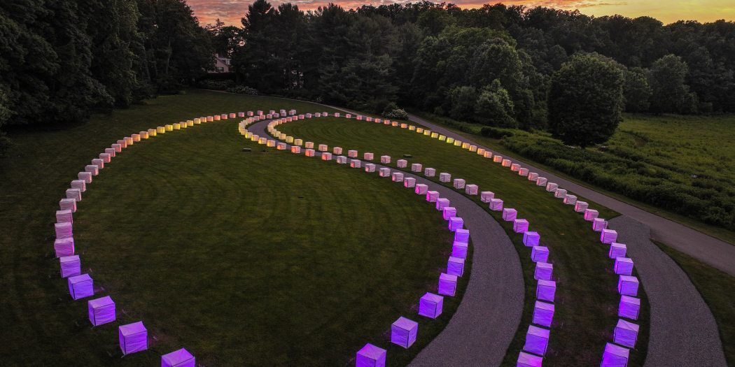 a series of cubes lit with the colors of dusk, on a darkening green lawn with a backdrop of trees against a sky at dusk