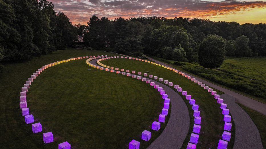 cubes lit in purple and gold, arranged in 2 long loops on a green lawn, seem to reflect the colors of dusk
