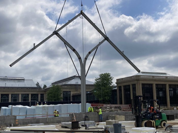 A large structure suspended by cables; 2 vertical steel beams split into a "Y" to support 2 more beams that form a peak pointing up to a cloud-filled sky