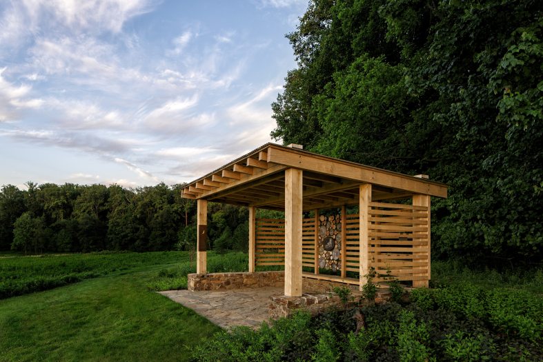 A wooden pavilion sits in a deep green open meadow with a small wood along the edge