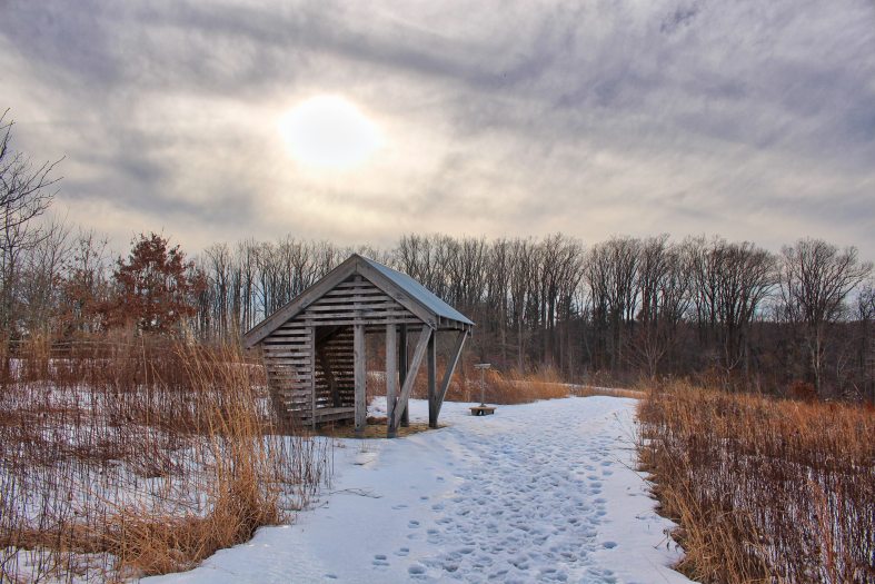 A small wooden structure stands along a snow-covered path in a large meadow