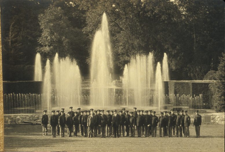 A group of men in military uniforms pose in a group picture in front of tall fountain jets in a garden