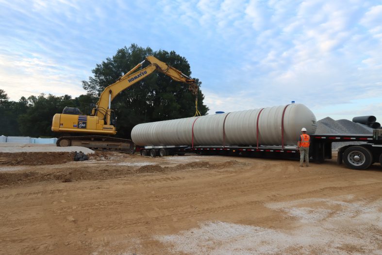 a large tank strapped to the flatbed of a truck, with a yellow crane in the background and a worker in an orange vest and white hardhat in the foreground