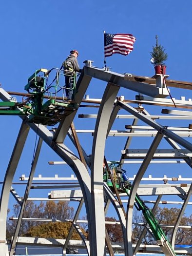 a construction worker stands in a bucket lift at the top of arched steel beams, decorated by an American flag and a Christmas tree