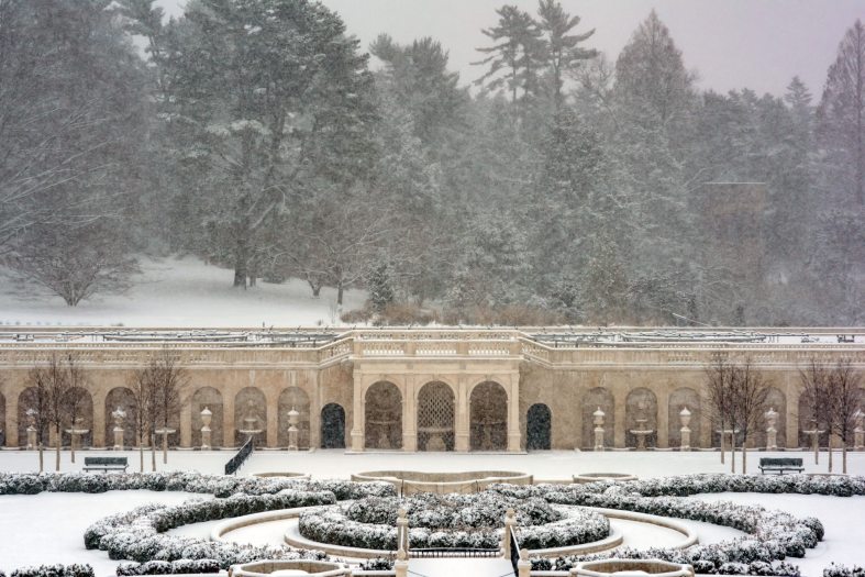 snow falls and covers the ground on a grand view of old-growth trees, clipped boxwood bushes, and a classical stone façade