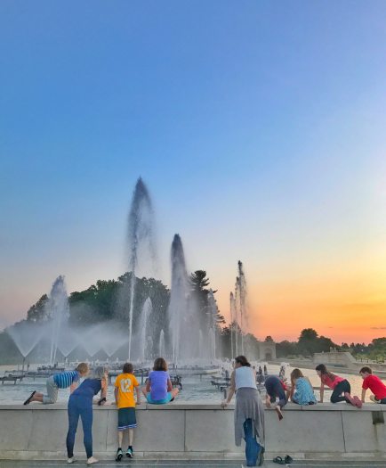 In front of a bright blue and vivid orange sunset, adults and children peer over a short stone wall into a rectangular fountain basic with jets shooting into the air.