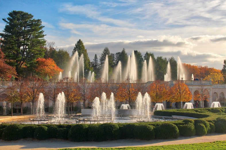 the low, golden fall sun illuminates sparkling fountain jets set against a background of green conifers, deciduous trees with bright orange leaves, and a cloud-filled blue sky