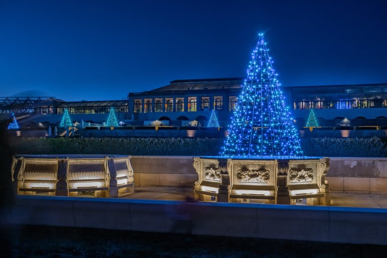 A view of carved limestone fountain basins adorned with Christmas lights in cone shapes, with a sprawling Conservatory in the background
