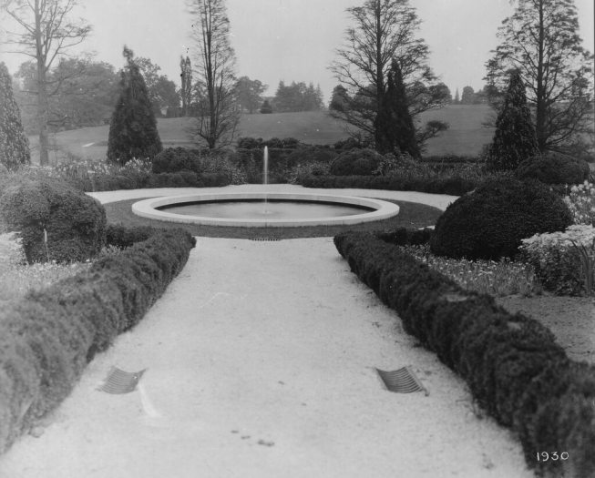 black and white photo of a boxwood-lined gravel pathway leading to a circular fountain with a single jet surrounded by a narrow ring of grass