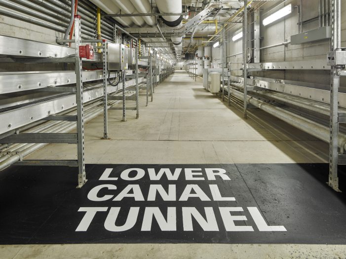 A long underground hallway lined with metal tubes is seen with the words "Lower Canal Tunnel" painted in black and white on the ground