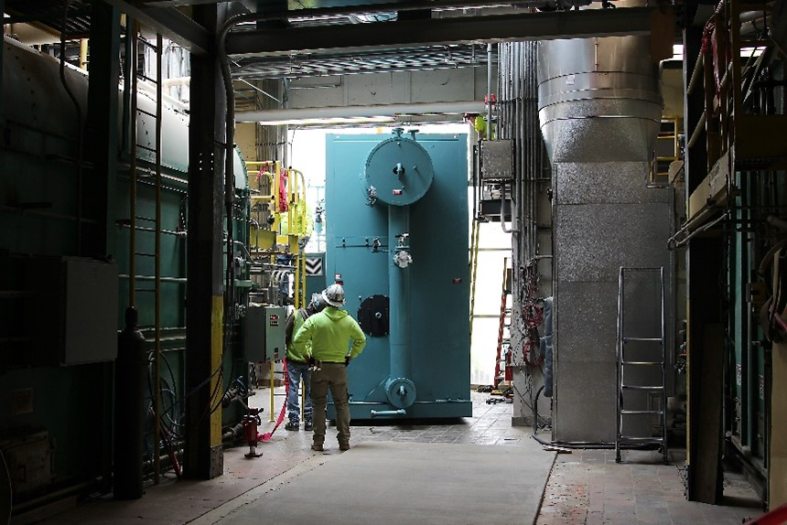 a person in a hard hat standing among large pipes and a teal boiler