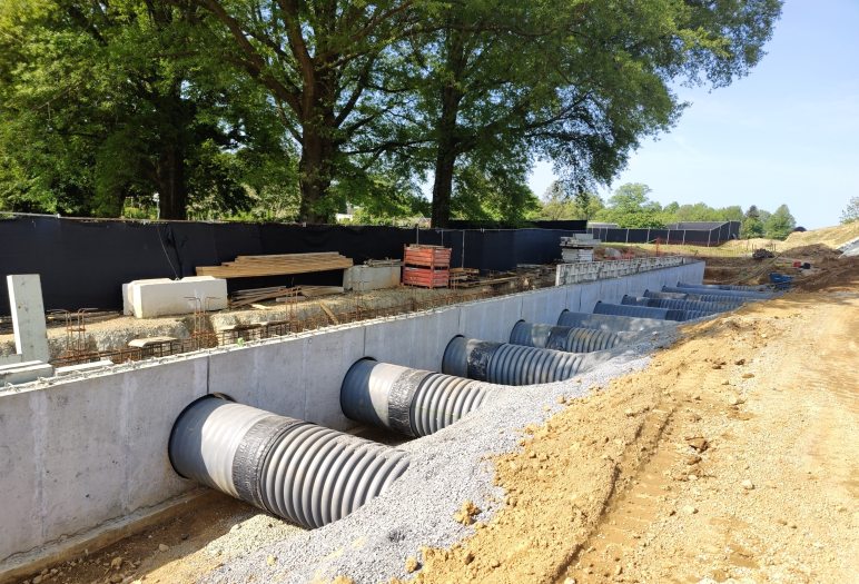 A series of 10 large ducts emerge from beneath dirt and gravel, then penetrate circular openings in a concrete wall just below ground level.