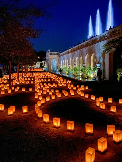 Luminaria glow in arching patterns on the ground, against a backdrop of a stone facade with illuminated fountain jets, pools, and plants.