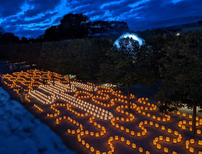 Thousands of gold and white luminaria in a pattern that resembles stitching glow against a dark blue sky and a fountain silhouette.