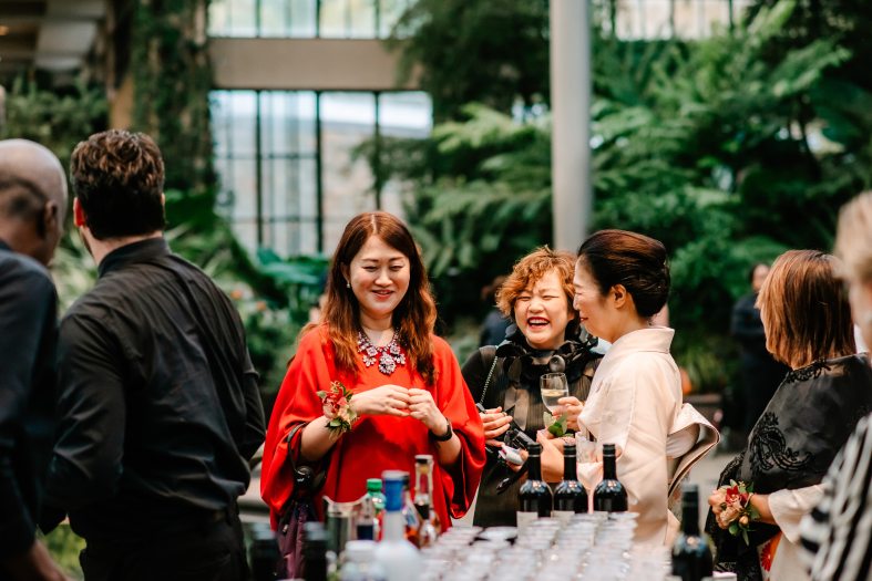 Closeup of people gathered in formal clothes in a conservatory setting, with focus on 3 people smiling and laughing, standing near a table filled with glasses and wine bottles.