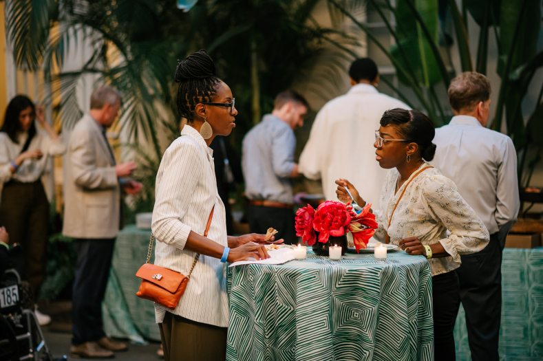 Focus on two people with appetizers in conversation at a tall table decorated with large red blooms in a conservatory setting, with other people helping themselves to food in the background.
