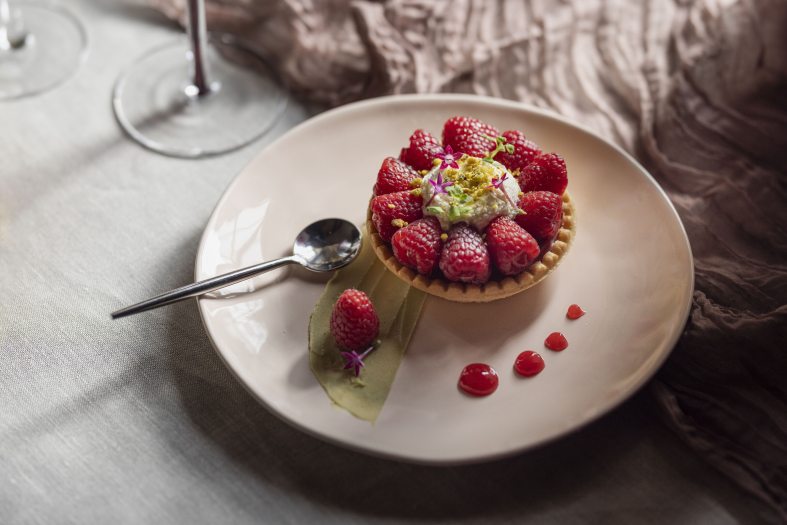 Closeup of a raspberry tart, along with a single raspberry and tiny purple flower on a leaf and dots of red gel artfully arranged on a plate with a spoon.