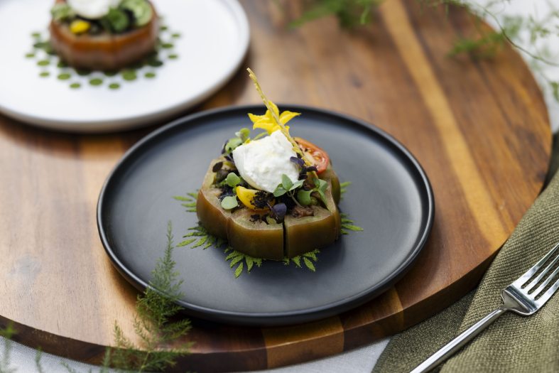 Closeup of a vegetable appetizer topped with white cream and a yellow flower on a round black plate.