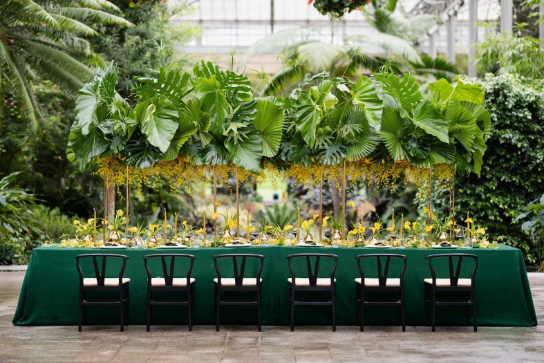Dining chairs and long table covered with dark green tablecloth, with individual place settings decorated with yellow calla lilies and a large overhead floral display of green palm leaves, ferns, and a variety of yellow flowers.