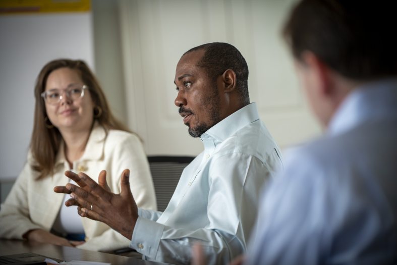 Closeup of three people sitting at a table in a conference room, two listening while one speaks.