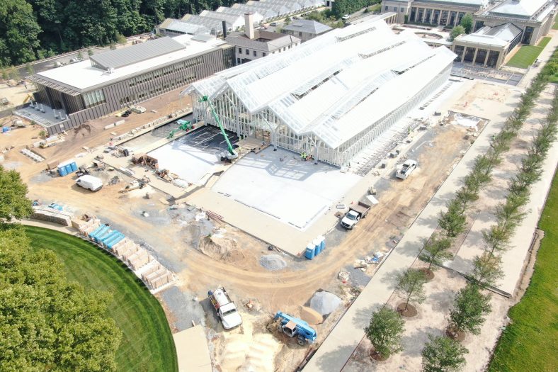 A large conservatory under construction, with a long ceiling of four rows of arched glass, new plantings of trees to the outside right, an empty waterlily courtyard and older conservatory in the background, and a large office building, boiler room, and production greenhouses in a long row of glass triangles at the left of the image.